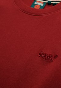 Camiseta de algodón burdeos con cuello redondo, que presenta un bordado texturizado de "SuperDry" en el área del lado izquierdo del pecho.