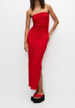 Femme portant une robe midi rouge texturée sans bretelles avec une fente latérale, associée à des sandales à plateforme rouges avec semelles noires.