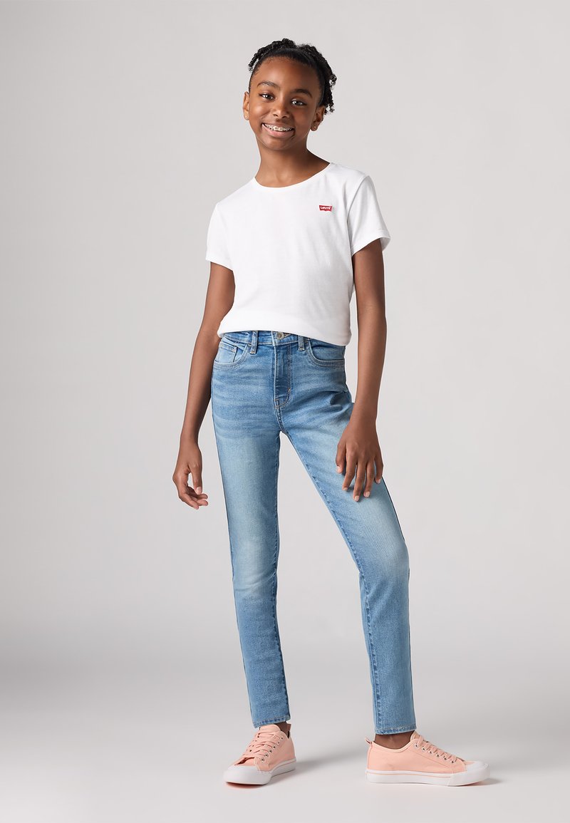 Camiseta blanca de manga corta con un pequeño logotipo rojo, combinada con jeans ajustados de color azul claro y zapatillas de deporte rosas. Corte recto y ajuste casual.