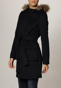 Manteau bleu marine foncé en mélange de laine, avec une capuche en fausse fourrure, une taille ceinturée, deux poches avant et un design épuré.