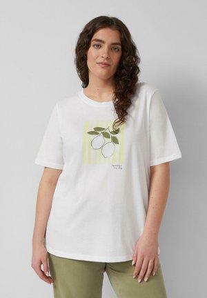 Kobieta o kręconych włosach w białej koszulce z grafiką cytryny i napisem "wyciśnij dzień", w połączeniu z jasnooliwkowymi spodniami.