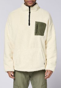Off-white Fleece-Pullover mit schwarzem Reißverschluss, grüner quadratischer Tasche mit Text und Kängurutasche an der Vorderseite.