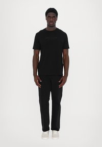 Schwarzes T-Shirt mit kurzen Ärmeln und dezentem Logo-Design, kombiniert mit schwarzen Hosen und hellen Turnschuhen. Glatte Stoffqualität und lässige Passform.