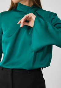 Turkoosi paita, jossa on korkea kaula-aukko ja leveät hihat, näyttäen sileältä ja minimalistiselta. Yhdistetty mustiin housuihin.