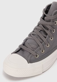 Gray canvas high-top sneaker with a white rubber toe cap, metallic eyelets, and textured detailing along the sole and side.