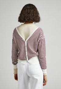 Chemise à manches longues rayée rouge et blanc avec un dos en V, ornée de boutons et en tissu texturé. Portée avec un pantalon blanc.