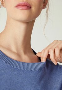 Pull bleu en maille côtelée avec un large décolleté ; gros plan sur la zone du cou et de l'épaule, mettant en valeur la texture douce et les coutures subtiles.