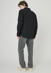 Un jeune homme aux cheveux bouclés se tient de dos, portant une chemise noire à manches longues, un jean gris et des chaussures épaisses blanc cassé sur un fond unicolore.