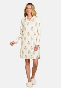 Vrouw in een crèmekleurige nachthemdjurk met bloemen en knopen, staand en met witte fluffy pantoffels tegen een effen achtergrond.