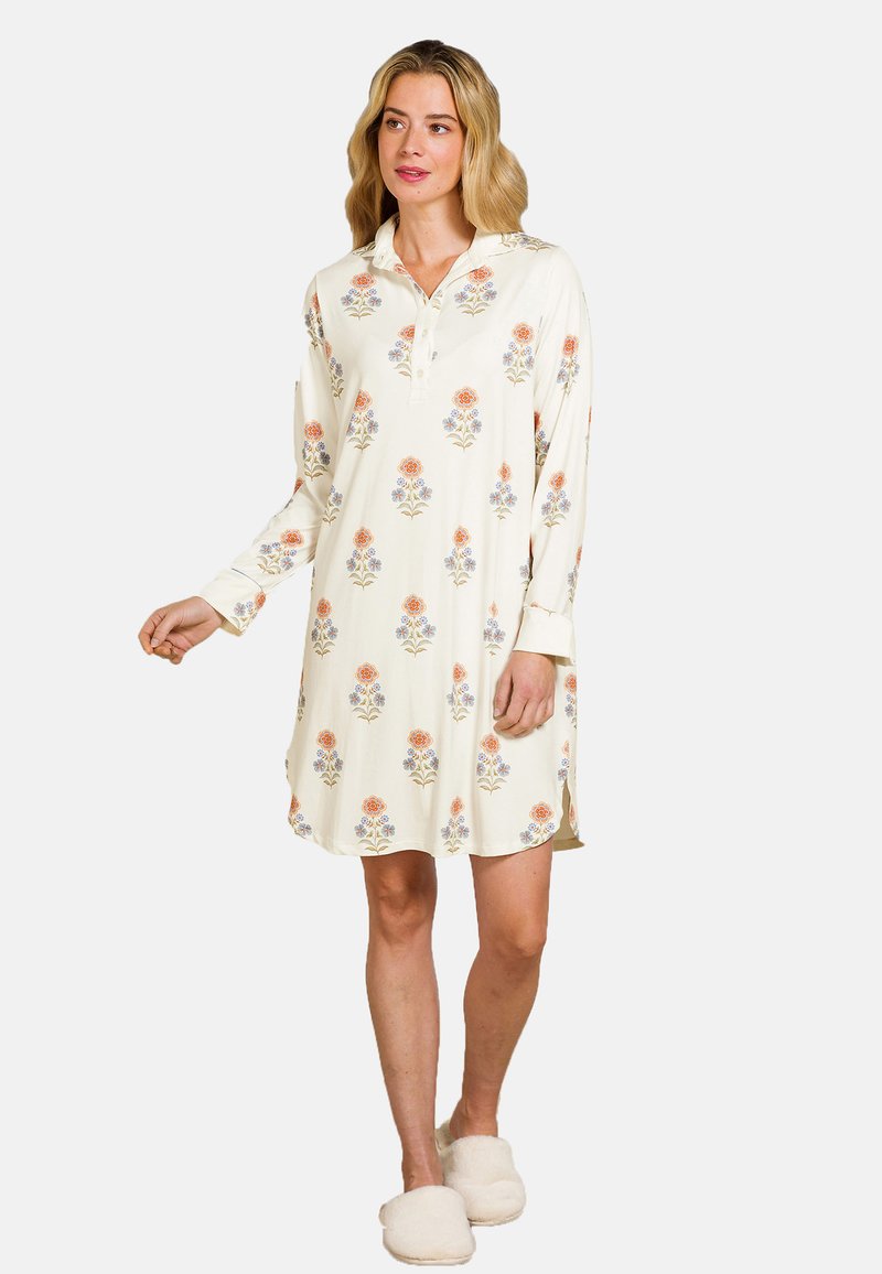 Vrouw in een crèmekleurige nachthemdjurk met bloemen en knopen, staand en met witte fluffy pantoffels tegen een effen achtergrond.