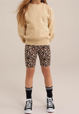 Enfant portant un sweat-shirt beige, un short imprimé léopard, des chaussettes noires avec des rayures blanches et des baskets montantes noires sur fond uni.