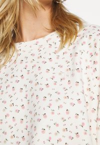 Bílá mikina s květinovým potiskem obsahující malé růžové a krémové květy, měkkou texturu a žebrovaný výstřih s jemným logem.