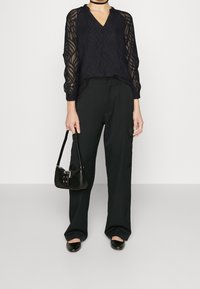 Blouse noire avec un motif ondulé texturé, encolure en V et manches longues ; associée à un pantalon cargo noir et un petit sac à main noir.