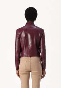 Giacca in pelle bordeaux con un design aderente, colletto alto e polsini con zip, abbinata a pantaloni beige, vista da dietro.