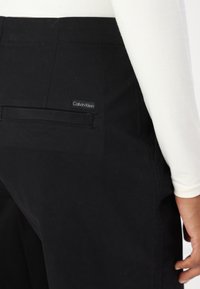Crne hlače glatke teksture, s stražnjim džepom i malom Calvin Klein oznakom. Dizajn je krojen s čistim linijama.