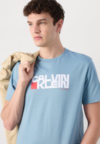 Svetlainio mėlynumo medvilnės marškinėliai su balta 0 CALVIN KLEIN1 logotipu, papuoštu raudona akcentu. Modelis laiko smėlio spalvos švarką per petį.