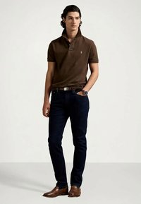 Ung man som bär en mörkbrun polo, mörka jeans, bruna läderskor och en armbandsur, stående med ena handen i fickan.