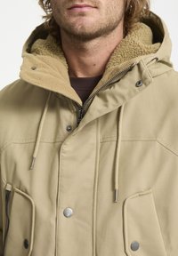 Beige jacket with a fleece-lined hood, snap and zip closures, two front pockets, and adjustable drawstrings. Smooth fabric with a soft texture.