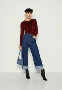 Haut long à manches longues bordeaux avec une texture, associé à un jean en denim taille haute à jambes larges avec des ourlets roulés. Le modèle porte un sac bleu.