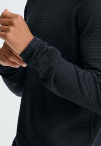 Donkergrijs shirt met lange mouwen en een geribbelde textuur. Voorzien van aangesloten manchetten en een ontspannen pasvorm, waarbij de structuur van de stof duidelijk zichtbaar is.