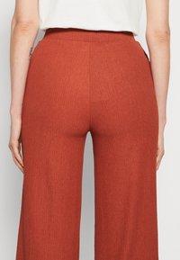 Vue de dos d'une personne portant un pantalon côtelé couleur rouille à taille haute, large aux jambes, et un haut blanc rentré dans le pantalon.