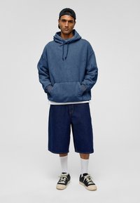 Blauwe oversized hoodie met een voorzak en trekkoord, gecombineerd met donkere denim shorts. Een wit t-shirt eronder, witte sokken en zwarte schoenen.