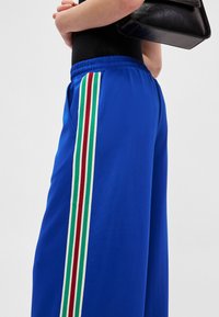 Μπλε αθλητικό παντελόνι με κορδόνι στη μέση, με κάθετες ρίγες σε πράσινο, άσπρο και κόκκινο κατά μήκος της πλευράς. Ομαλή υφή υφάσματος.
