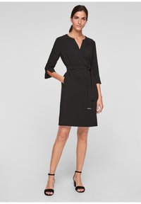 Robe noire enveloppante avec des manches trois-quarts, décolleté en V, ceinture à nouer à la taille et poches latérales. Tissu lisse avec une coupe décontractée.