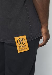 Juodas marškinėlių su stačiakampiu oranžiniu etikete, kurioje rašoma "240gsm" ir "pamesti veidai", pritvirtintu prie apatinių siūlių, yra matoma tekstūra.