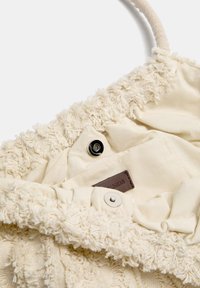 Beige fabric bag with textured, fluffy exterior, snap button closure, and a brown leather tag inside. Features a soft, ruffled design.