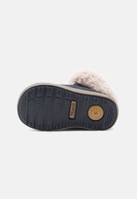 Semelle de botte avec surface en caoutchouc texturé, de couleur bleu marine foncé, ornée d'une bordure en fourrure beige et d'un accent circulaire de marque avec des motifs antidérapants.
