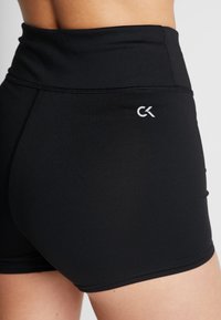 Sorte højtaljede atletiske shorts med et lille hvidt "CK" logo på højre hofte, iført af en person mod en ensfarvet baggrund.