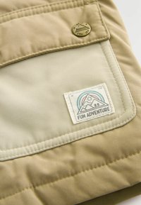 Tessuto outdoor beige con una patch cucita che presenta un design di montagna e sole. Include un bottone a pressione in ottone e una texture morbida.
