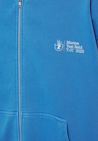 Sweat à capuche zippé bleu avec logo corail blanc et texte « Marine Test field T-07 2025 » sur la poitrine gauche au-dessus de la poche avant.