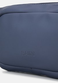 Ναυτικό μπλε αδιάβροχη τσάντα με λεία υφή, στρογγυλεμένες γωνίες και ανάγλυφο λογότυπο "RAINS". Ορατή λεπτομέρεια ραφής κατά μήκος της πλευράς.