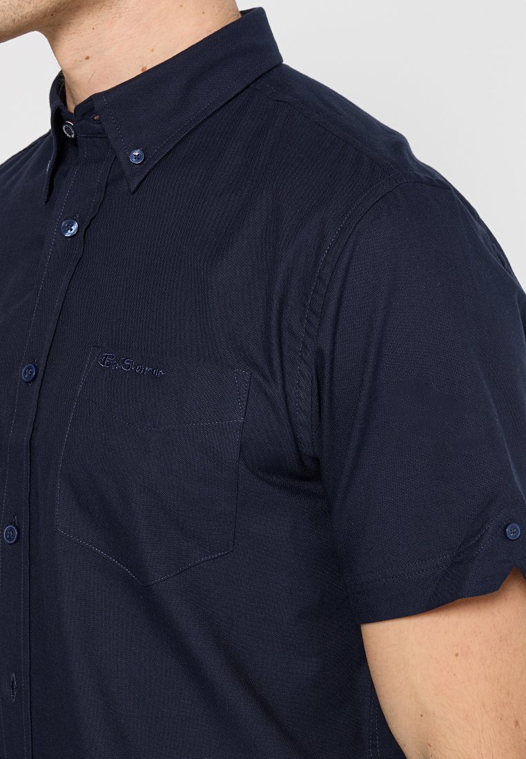 Homme portant une chemise à manches courtes bleu marine, avec une poche sur la poitrine et un col boutonné, visible de l'épaule au cou.