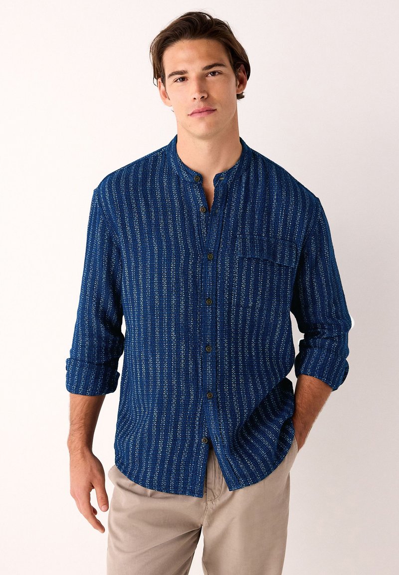 Jonge man met bruin haar, gekleed in een blauw overhemd met patroon en opgerolde mouwen, en beige broek, staand tegen een effen achtergrond.