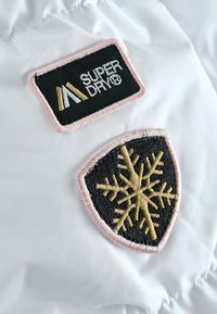 Tessuto bianco con due toppe ricamate. La toppa superiore riporta "SUPER DRY" in nero e oro su sfondo rosa chiaro. La toppa inferiore mostra un fiocco di neve dorato.