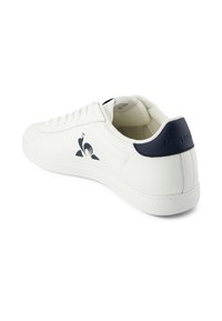 Sneaker in pelle bianca con un accento blu navy sul tallone, un logo sul lato, lacci piatti e una suola testurizzata.