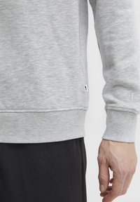Grå sweatshirt med ribbede manchetter og kant, lavet af et struktureret stof. Lille logo på sidesømmen og tommelhul-detalje på ærmet.