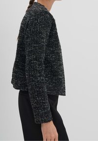 Veste en tweed noir avec une finition texturée, coupe courte et manches longues. Présente un col rond et de subtils accents métalliques.