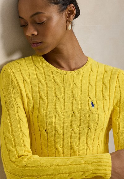 Κίτρινο πλεκτό πουλόβερ με σχήμα καλωδίου, γύρος λαιμός και ριμπ μανσέτες· μικρό μπλε λογότυπο στο στήθος. Απαλή υφή, εφαρμοστή γραμμή.