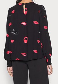 Blusa nera con maniche lunghe, caratterizzata da una stampa ripetuta di labbra rosse e testo bianco che recita "la vita è fantastica", dettaglio con apertura a chiave sul retro.