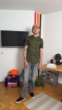 Camicia a maniche corte verde oliva con colletto rotondo, abbinata a jeans grigi chiari e sneakers nere. Tatuaggi visibili sulle braccia.