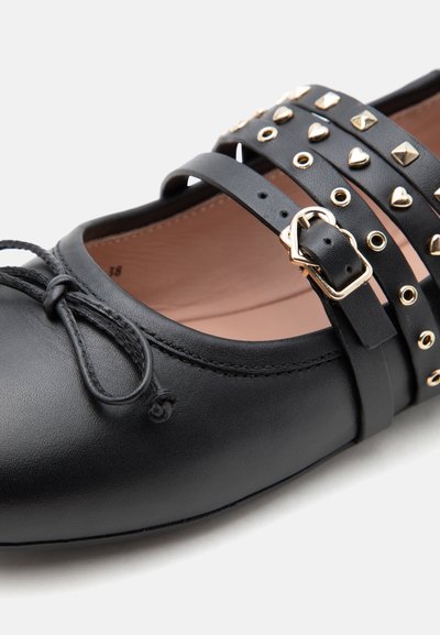 Zapato plano de cuero negro con detalle de lazo en la punta y tres tiras con tachuelas y hebillas doradas en la parte superior.