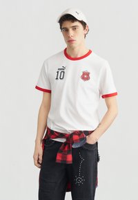 Νεαρός άνδρας που φοράει λευκό καπέλο Puma, λευκό πουκάμισο με κόκκινη επένδυση και αριθμό 10, κόκκινο πουκάμισο καρό δεμένο στη μέση και μαύρα τζιν με τρουκς.