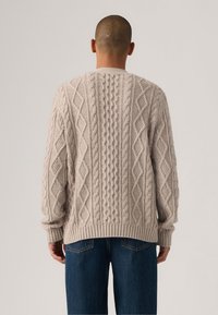 Man wearing beige thick cable-knit sweater and blue jeans, standing facing away against plain light background.