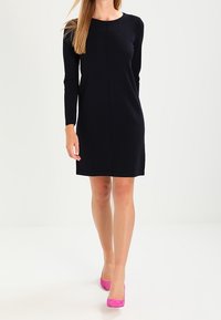 Schwarzes langärmliges Kleid mit geradem Schnitt und Rundhalsausschnitt. Getragen mit pinken Wildlederschuhen mit hohen Absätzen. Glatte Textur.