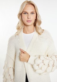 Krémový pletený kardigan s texturovanými rukávy, který se vyznačuje vystouplými vzory a otevřenými stehenními akcenty. Oversized střih a uvolněný design.