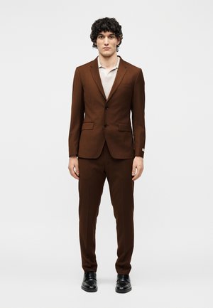 Mężczyzna o kręconych ciemnych włosach, ubrany w brązowy garnitur, czarne buty i jasnobeżową koszulę z kołnierzykiem, stojący na jednolitym tle.